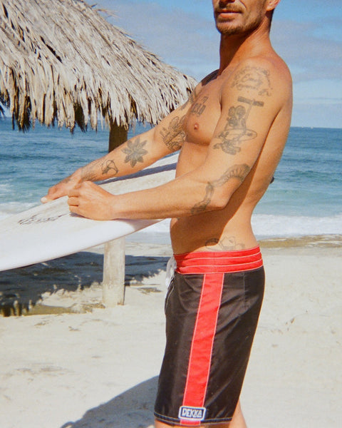 A shirtless, tattooed man holds a surfboard on a sandy beach. He wears 310 Dekka - Black/Red boardshorts. A thatched hut and the ocean appear in the background beneath a clear blue sky.