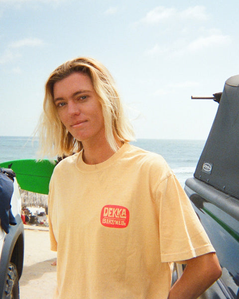 A person with shoulder-length blonde hair wears a Birdwell x Dekka T-Shirt in mustard on a beach near parked vehicles, with the ocean and a partly cloudy sky in the background.