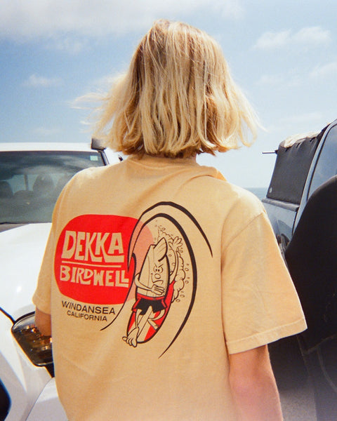 A blonde-haired person wears a mustard Birdwell x Dekka T-Shirt, made from 100% garment-dyed cotton with a bold graphic and 