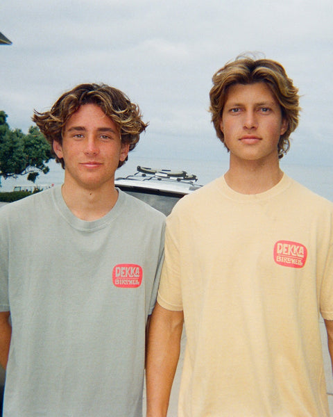 Two young men with wavy hair stand outdoors, both wearing the Birdwell x Dekka T-Shirt in Sandstone. A parked car and trees are visible in the background.
