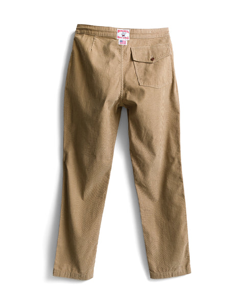 Introducing the Classic Corduroy Pants - Sand: crafted in Southern California from 100% cotton corduroy, these tan pants are pictured laid flat from the back. They feature two rear pockets—one secured with a button—and are finished with a distinctive white and red label at the waistband.