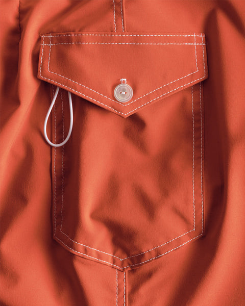 Close-up of the paprika-colored fabric side pocket from the 808 SurfStretch Side Pocket Boardshorts, featuring white stitching, a round button, and a white loop partially tucked under the pocket flap—details that showcase the premium craftsmanship of these USA-made boardshorts.