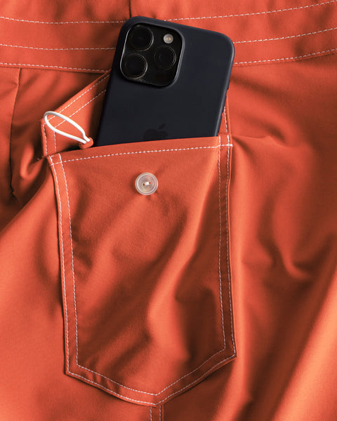 A black smartphone is partially tucked into the front pocket of the 808 SurfStretch Side Pocket Boardshorts in Paprika, with white stitching and a button closure visible.