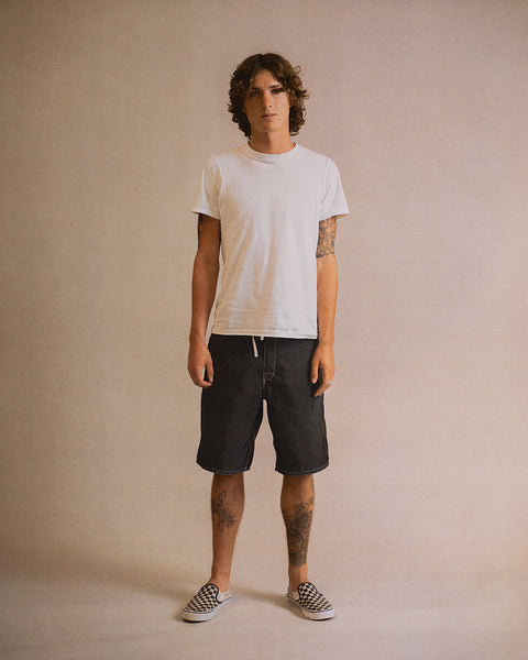 A person with curly hair stands facing forward, wearing a white t-shirt, the 312 Boardshorts - Black, and checkered slip-on shoes. Their arms and legs display visible tattoos. The background is plain and neutral.