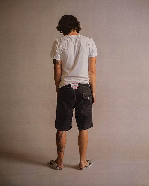 A person with curly hair, wearing a white t-shirt, 312 Boardshorts - Black made in the USA, and patterned slip-on shoes stands facing away from the camera against a plain background.