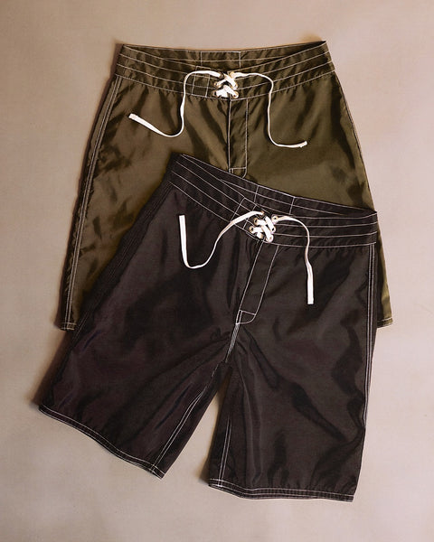Two pairs of 312 Boardshorts in black and dark olive, made in the USA, are laid out on a neutral background, slightly overlapping. These long-length boardshorts feature visible white stitching and white drawstrings for a classic look.