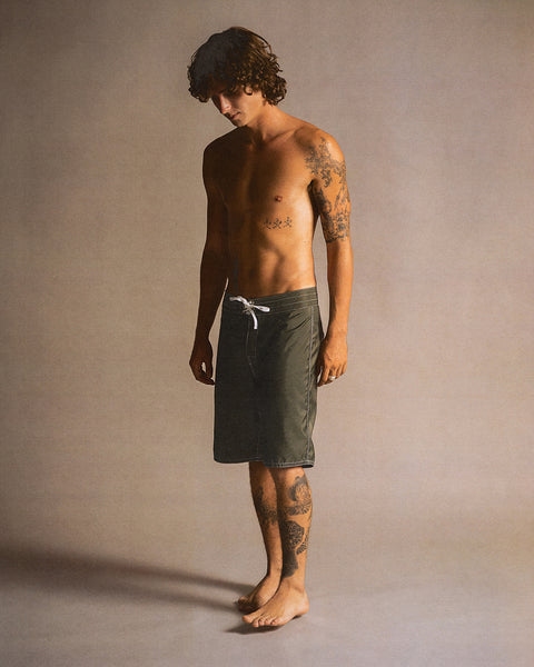 A shirtless person with curly hair and many tattoos stands barefoot, wearing knee-length 312 Boardshorts - Dark Olive (made in USA SurfNyl), and looks down against a neutral background.