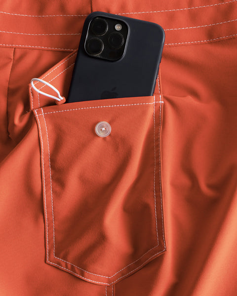 A black smartphone partially tucked into the buttoned pocket of orange 806 SurfStretch Side Pocket Boardshorts in Paprika, featuring white stitching and made in the USA.
