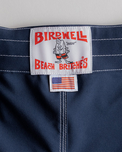 A close-up of the navy waistband on the 806 SurfStretch Side Pocket Boardshorts, featuring a Birdwell Beach Britches label with a cartoon bird and a small American flag tag below it—highlighting these classic stretch boardshorts proudly made in the USA.