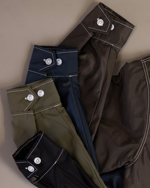 Three Racing Jacket 26 shirts in Dark Olive, Bone, and Black are neatly folded on a neutral background, each featuring white stitching and buttoned cuffs inspired by Birdwell’s classic SurfNyl craftsmanship.