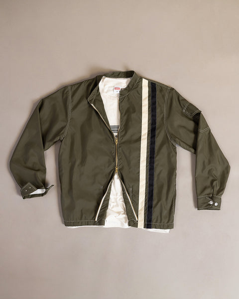 The Racing Jacket 26 in Dark Olive/Bone/Black, featuring beige and black vertical stripes on the right side and inspired by classic Birdwell SurfNyl designs, is shown laid flat over a white t-shirt on a neutral background.