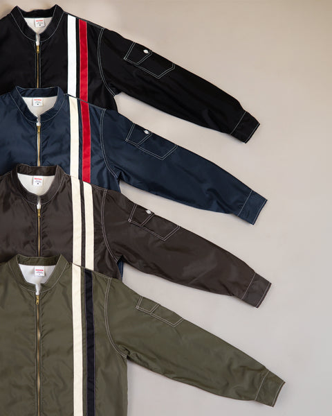 Four Racing Jackets 26 in black, navy, brown, and dark olive/bone/black are laid flat. Each features SurfNyl construction with a vertical stripe on the left, a sleeve pocket, and a gold zipper. Set against a neutral grey-beige background—a signature Birdwell touch for timeless style.