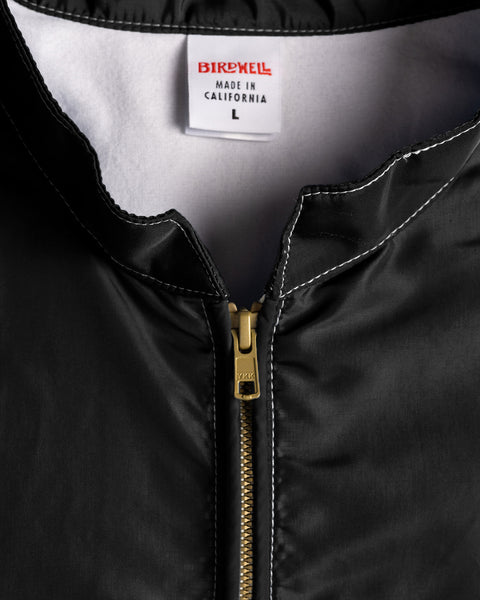 Close-up of the Racing Jacket 26 in black with white and red accents, featuring a vintage-inspired design with a gold zipper and visible white stitching. The label inside reads BIRDWELL MADE IN CALIFORNIA L, emphasizing its American-made quality. The inner lining is white.
