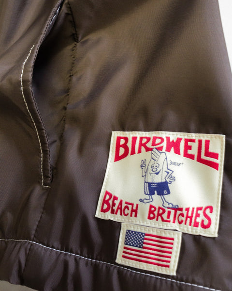 Close-up of brown Racing Jacket 26 - Brown/Bone SurfNyl fabric with a Birdwell Beach Britches patch featuring a cartoon bird, bold red text, and a small American flag label underneath. Part of a pocket seam is visible on the left, showcasing the iconic American-made craftsmanship.