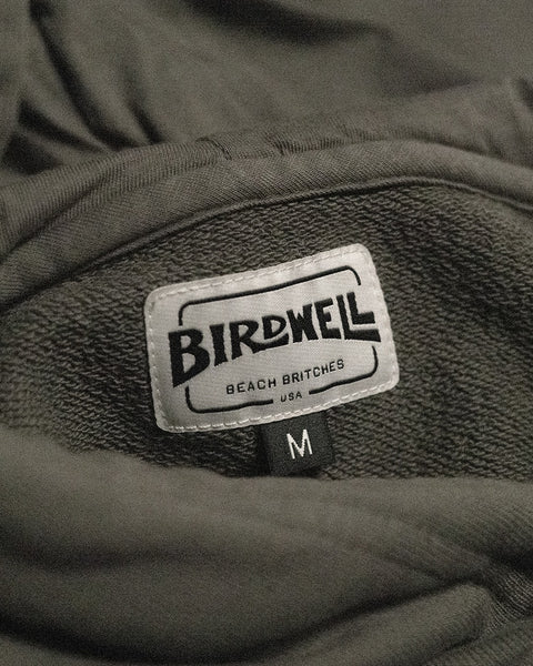 A close-up of the Harmony Hoodie - Dark Olive’s label, reading Birdwell Beach Britches USA and sewn onto dark gray French terry fabric, with a black size tag marked M beneath it—highlighting classic, made-in-USA craftsmanship reminiscent of a premium hoodie.