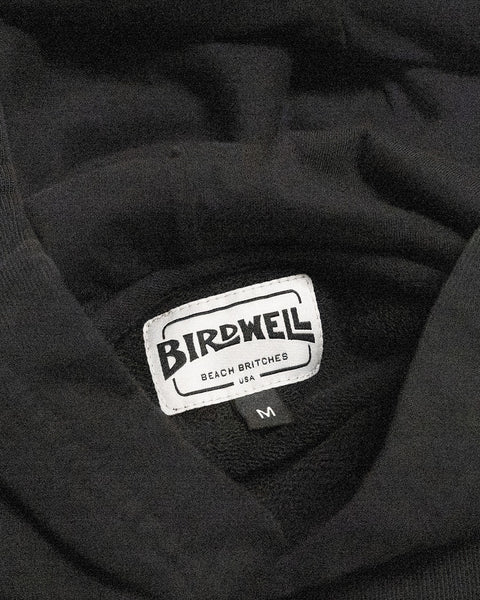 A close-up of the Harmony Hoodie in Faded Black shows a label that reads Birdwell Beach Britches USA, highlighting its made-in-USA origin, along with a small tag below marked with an M to indicate the medium size.