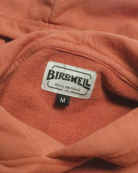 A close-up of a clothing label on an orange Harmony Hoodie - Paprika highlights its made-in-USA craftsmanship with “BIRDWELL BEACH BRITCHES USA” text, and a black tag below displaying the size M.