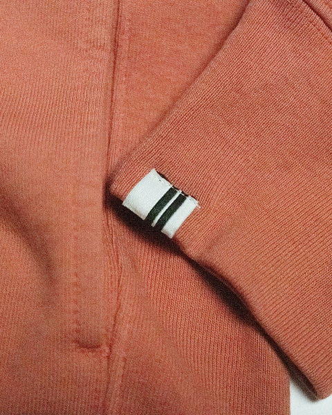 A close-up of the Harmony Hoodie - Paprika shows its pinkish-orange French terry sleeve with a white tag featuring two black stripes, resting on matching fabric.