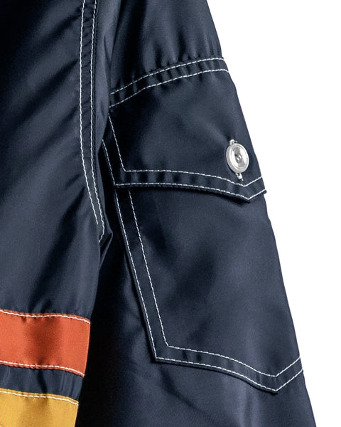 Close-up of a navy blue Comp Jacket 26 - Navy/Gold/Paprika sleeve, featuring white stitching, a flap pocket with a silver snap button, and horizontal gold and paprika stripes near the cuff—crafted with American-made precision and SurfNyl durability.