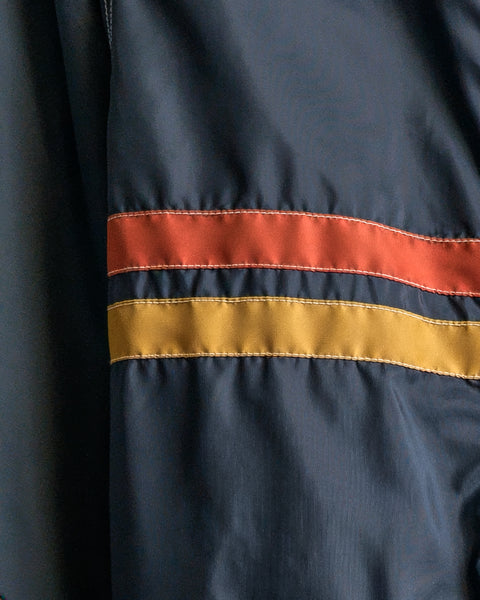 A close-up of the Comp Jacket 26 - Navy/Gold/Paprika reveals dark blue fabric with bold gold and paprika horizontal stripes stitched across it, highlighting the celebrated SurfNyl durability found in every American-made Birdwell jacket.