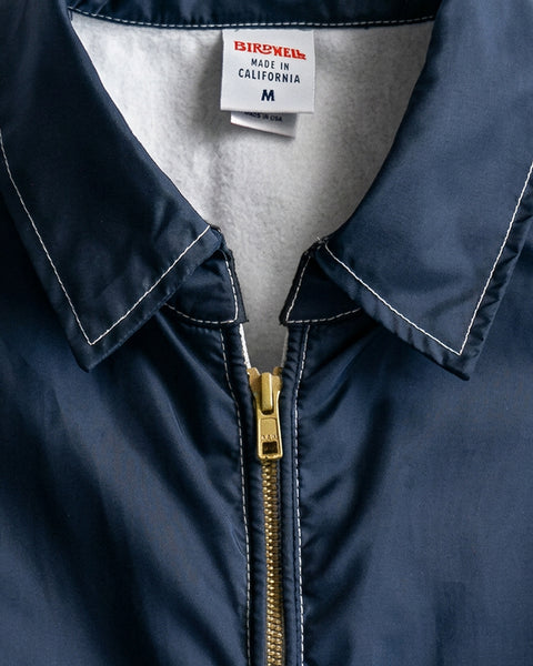 Close-up of the Comp Jacket 26 - Navy/Gold/Paprika, featuring navy blue SurfNyl fabric with white stitching, a gold zipper, and a visible tag that reads BIRDWELL MADE IN CALIFORNIA M. The jacket includes a white inner lining and exemplifies classic American-made craftsmanship and Birdwell’s renowned durability.