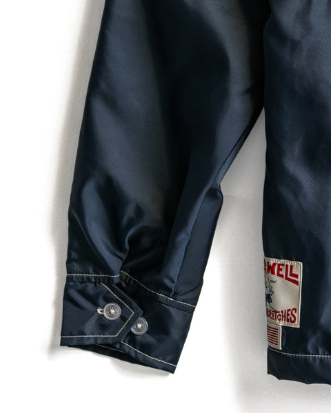 A close-up of the Comp Jacket 26 - Navy/Gold/Paprika sleeve reveals a buttoned cuff and a stitched white patch with red text near the hem, highlighting classic American-made craftsmanship and SurfNyl durability.