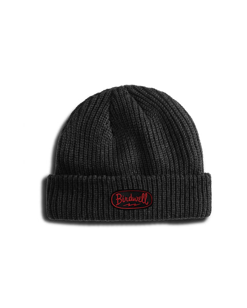 The Powerbird Watchcap - Coal is a black knit beanie featuring a wide ribbed design, a folded cuff, and a red and white Powerbird patch on the front, shown against a white background.