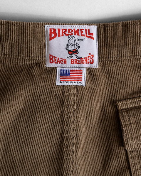Close-up of the waistband on the Classic Corduroy Shorts - Sand, crafted from 100% cotton corduroy in a warm brown shade. Two sewn labels are featured: one displaying Birdwell Beach Britches with a cartoon bird, and another reading Made in U.S.A. with an American flag. The adjustable drawcord waist provides a comfortable, personalized fit.