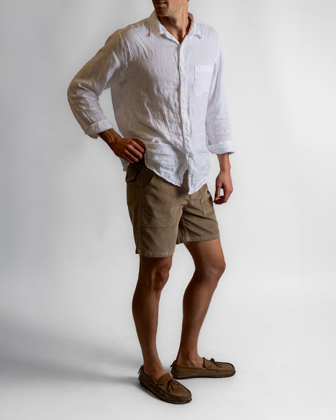 A person stands against a plain background wearing a white long-sleeve button-up shirt, the Classic Corduroy Shorts in Sand with an adjustable drawcord waist, and brown loafers. The person’s head is slightly turned, and one hand rests on their hip.