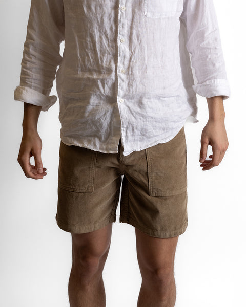 A person wearing Classic Corduroy Shorts - Sand featuring an adjustable drawcord waist and a white long-sleeve button-up shirt stands against a plain white background, with their hands relaxed at their sides.