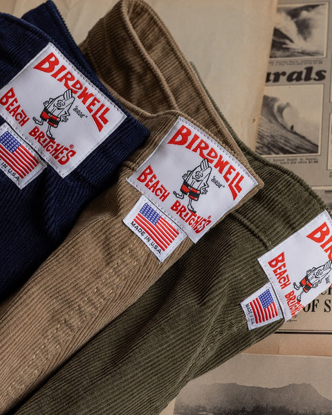 Three pairs of Classic Corduroy Shorts - Sand in navy, tan, and olive—each crafted from 100% cotton—feature a Birdwell Beach Britches patch and a Made in USA label with an American flag. Designed with an adjustable drawcord waist for the perfect fit, they are laid out on vintage papers.