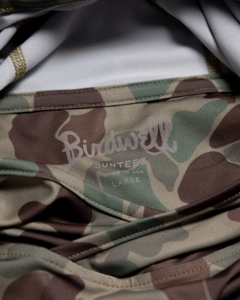 Close-up of a clothing label on Frogskin Camo fabric, showing the Birdwell brand name, the word Surf, and the size Large. The label also highlights that the Sunteez Hoodie is UPF50+ and made in USA.