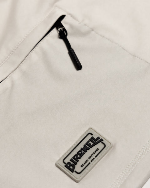 Close-up of a light-colored fabric pocket with a black zipper and a rectangular Birdwell Beach Britches logo patch sewn below it, crafted from moisture-wicking fabric. The Sunteez Hoodie - Bone offers UPF 50+ sun protection and is proudly made in the USA, blending comfort and protection in every stitch.