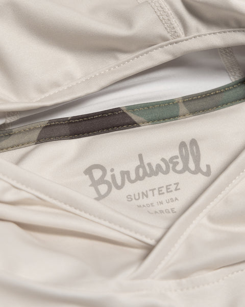 Close-up of the Sunteez Hoodie - Bone, crafted from moisture-wicking fabric. The inner label reads “Birdwell SUNTEEZ Made in USA Large,” and a section of camouflage-patterned waistband is visible above the label.