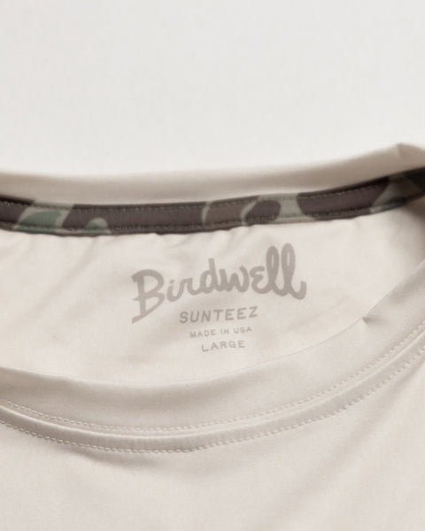 Close-up of the collar area of the Sunteez Short Sleeve - Bone t-shirt from Birdwell, featuring a printed label reading “Birdwell Sunteez Made in USA Large” and a camouflage-patterned inner collar lining. Crafted from performance stretch fabric, this shirt delivers flexibility and comfort for every adventure.