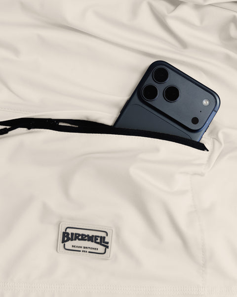 A black smartphone partially tucked into a zippered pocket of cream-colored Sunteez Short Sleeve - Bone shorts with a Birdwell Beach Britches label below the pocket, crafted from moisture-wicking, performance stretch fabric for all-day comfort.