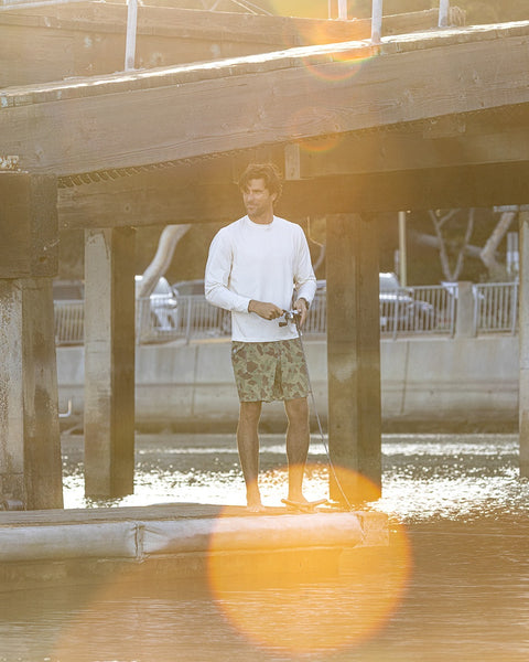 A person wearing a Sunteez Long Sleeve - Bone shirt and green shorts stands on a dock fishing under a wooden pier, with sunlight and lens flare effects in the image.