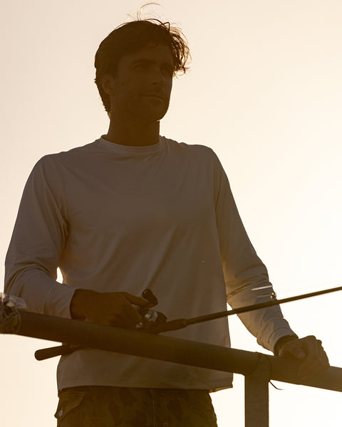 Wearing the Sunteez Long Sleeve - Bone, a man fishes at sunset behind a railing, holding his fishing rod, his face mostly silhouetted by the bright backlighting.