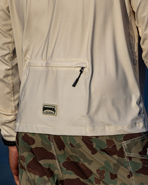 A person wearing the Sunteez Long Sleeve - Bone, featuring a light beige color with a zippered pocket and a patch on the back, paired with green and brown camouflage pants crafted from moisture-wicking fabric.