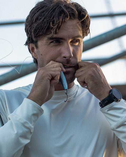 A man with tousled dark hair and a black wristwatch holds a fishing lure between his lips, both hands raised, while wearing the Sunteez Long Sleeve - Bone shirt. The background is blurred.