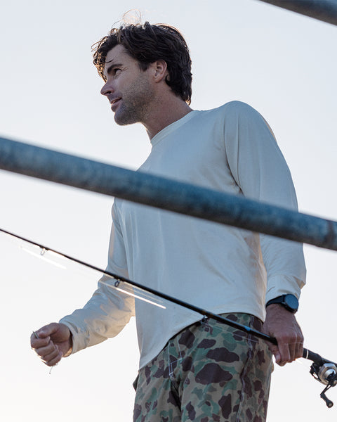 A man wearing the Sunteez Long Sleeve - Bone shirt and camouflage pants holds a fishing rod and looks to the side, with a watch on his wrist and metal bars visible in the foreground.