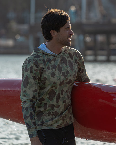 A man in the Sunteez Hoodie - Frogskin Camo stands by the water, holding a red kayak under his arm. Wearing this moisture-wicking UPF50+ shirt, he’s ready for any adventure. The background is blurred, with docks and boats faintly visible.