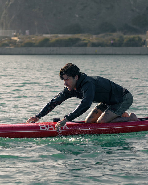 A person crouches on a red paddleboard labeled BAKA, balancing over calm water near a rocky shoreline with greenery and a concrete wall in the background, wearing the Sunteez Hoodie - Black for UPF50+ sun protection.