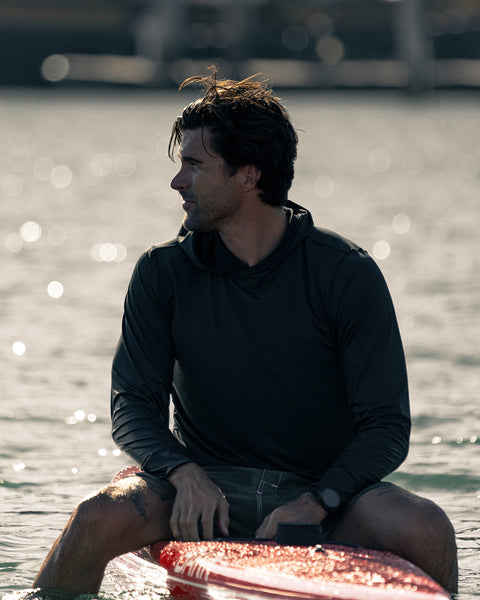 A man wearing the Sunteez Hoodie - Black sits on a red surfboard in calm water, looking to his left as sunlight reflects off the water behind him.