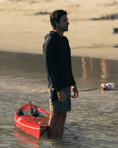 A man stands in shallow water next to a small red kayak on a sandy shore. He wears the Sunteez Hoodie - Black and shorts—his UPF50+ shirt provides extra protection as he faces left with a calm, thoughtful expression. The background features more beach and water.