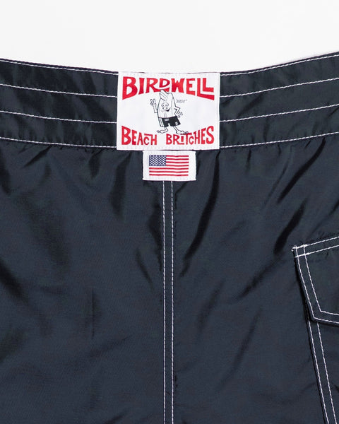Close-up of the 312 Boardshorts in navy, featuring white stitching, a Birdwell Beach Britches label, and a small American flag tag above the pocket—all highlighting their long-length style and authentic “made in the USA” craftsmanship.