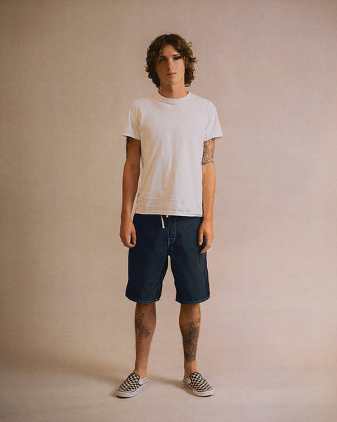 A person stands against a plain background, wearing a white t-shirt, navy 312 Boardshorts made in the USA that fall to the knees, and checkered slip-on shoes. They have curly hair, visible tattoos, and a neutral expression.