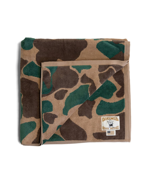 The Camo Towel - Frogskin is a folded, oversized towel featuring a camouflage pattern of green, brown, and tan shapes with a prominent Birdwell Beach Britches logo patch on the corner. Crafted from soft 100% cotton, it is proudly made in the USA.