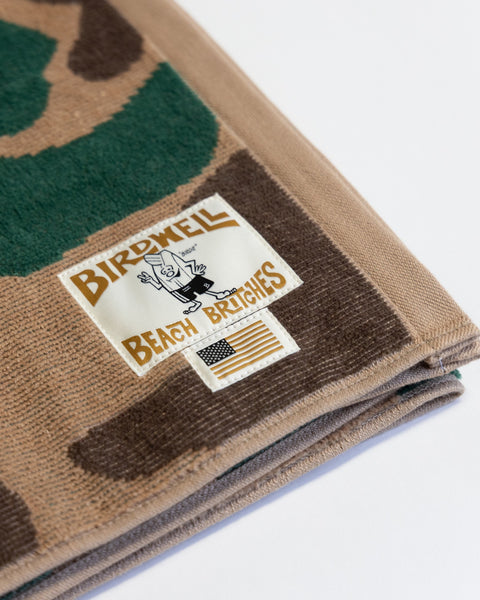 The Camo Towel - Frogskin is a folded, oversized towel with a brown and green camouflage pattern, featuring a Birdwell Beach Britches label and a small American flag tag. Made in the USA from 100% cotton towel material, it offers comfort and classic style.