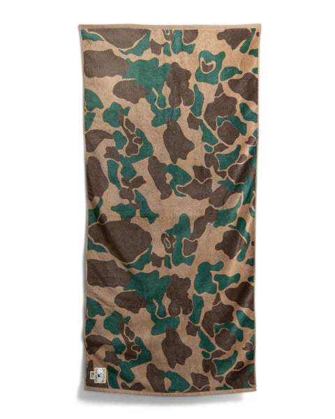 The Camo Towel - Frogskin, a rectangular oversized towel with a green, brown, and tan camouflage pattern, lies flat against a white background. Made from 100% cotton, it features a small white tag visible in the bottom left corner.